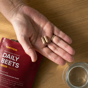 Older hand holding two Daily Beets capsules with the pouch and a glass of water on an oak counter